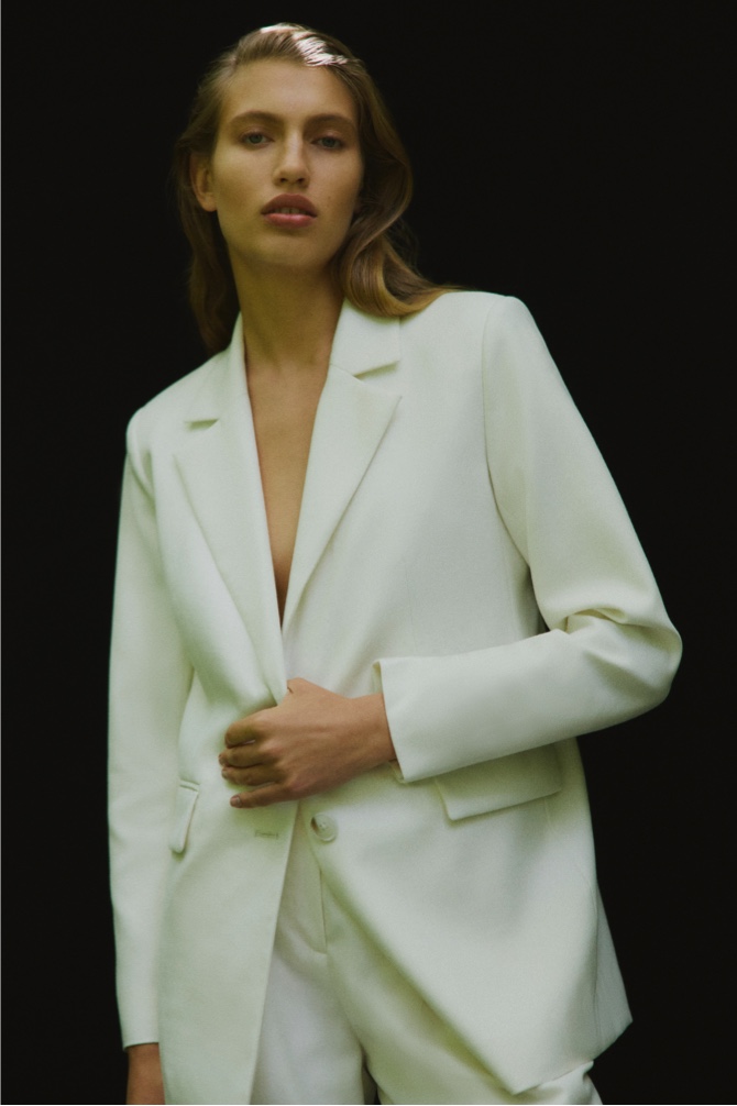 Woman in black wool blend double-breasted blazer coat, cream wool blend single-breasted boxy blazer, black cut out rib tank and cream wool blend relaxed rise trouser