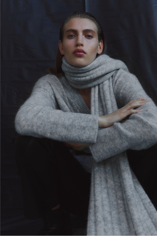 Woman in light grey marle tactile v neck knit, black leather straight leg pant, black twisted buckle belt and light grey marle wide rib knit scarf