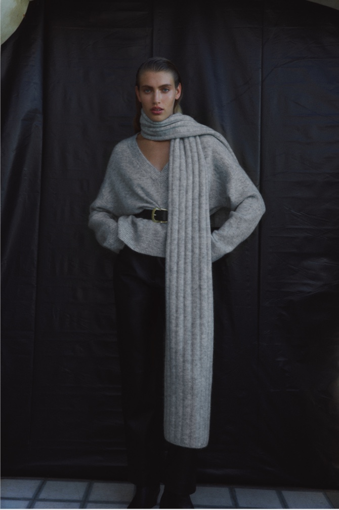 Woman in light grey marle tactile v neck knit, black leather straight leg pant, black twisted buckle belt and light grey marle wide rib knit scarf