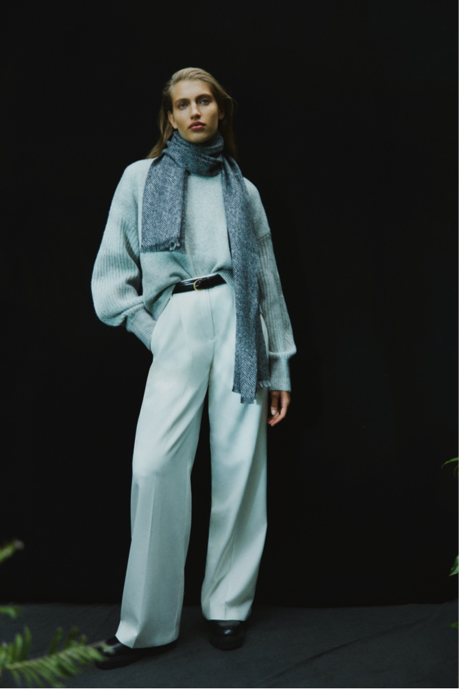 Woman in light grey marle stitch sleeve knit, cream wool blend relaxed rise trouser, black double wrap belt and black herringbone scarf