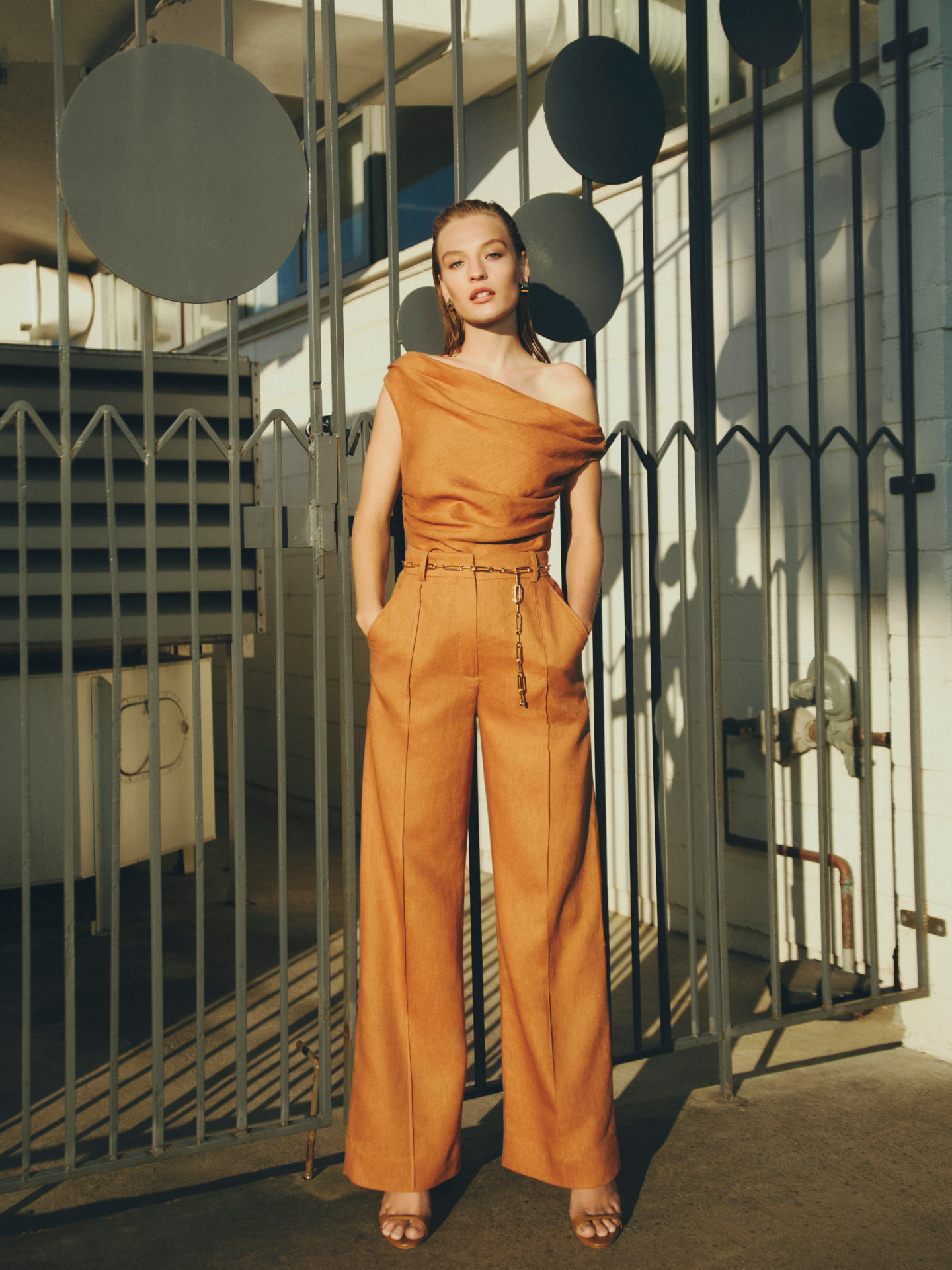 Woman in nutmeg off shoulder top, nutmeg high rise pants, gold elle chain belt, nutmeg isla wedge and gold lacey earrings